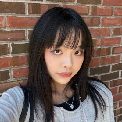 Xinyue Huang, a young Chinese poet, stands against a brick wall. She has long black hair, bangs that fall to her eyes, and is wearing a black shirt underneath a light grey shirt and a silver heart pendant on a chain.