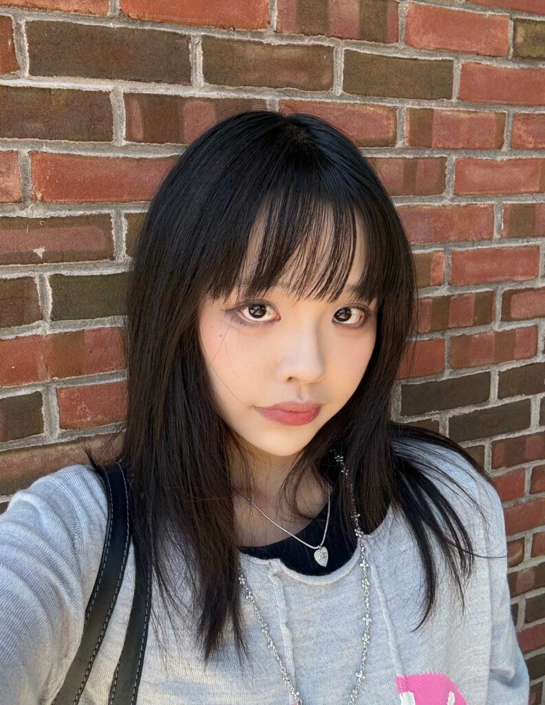 Xinyue Huang, a young Chinese poet, stands against a brick wall. She has long black hair, bangs that fall to her eyes, and is wearing a black shirt underneath a light grey shirt and a silver heart pendant on a chain.