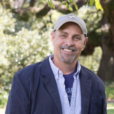 Jack Bedell, an older white male, wears a tan baseball cap, collared shirt, and blue sports coat. He is standing outside in the sun and smiling with oak trees in the background.