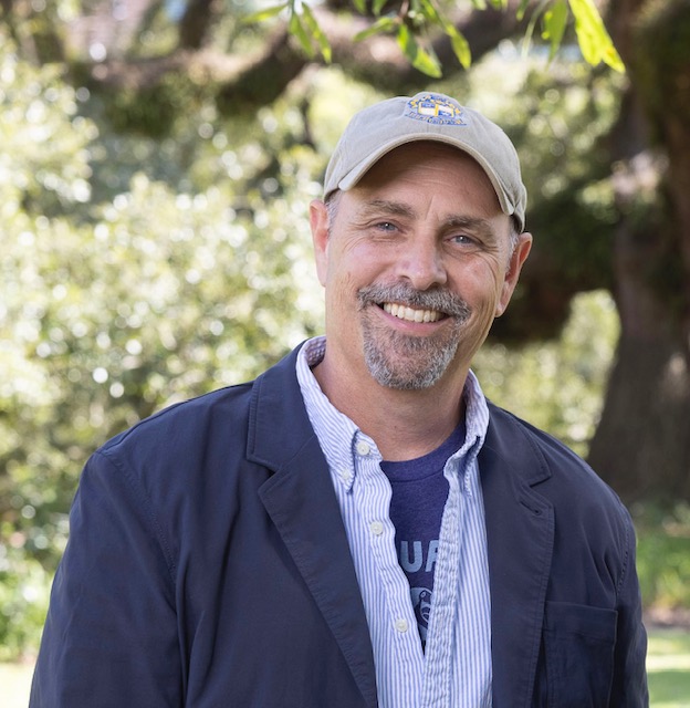 Jack Bedell, an older white male, wears a tan baseball cap, collared shirt, and blue sports coat. He is standing outside in the sun and smiling with oak trees in the background.