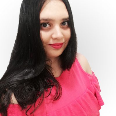 A South Asian woman with long black hair stands against a white wall. She has dark brown eyes and is wearing a bright pink top with her left shoulder exposed.