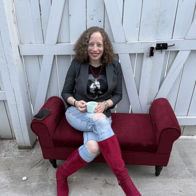 Francesca Leader, a fair-skinned woman with curly shoulder-length red hair, sits outdoors on a wine-red chaise longue in front of a white picket fence. She is wearing a black tshirt, pale blue jeans with holes in the knees, and red velvet, calf-high, zip-up boots. She is smiling widely and holding a small cyan mug with both hands.