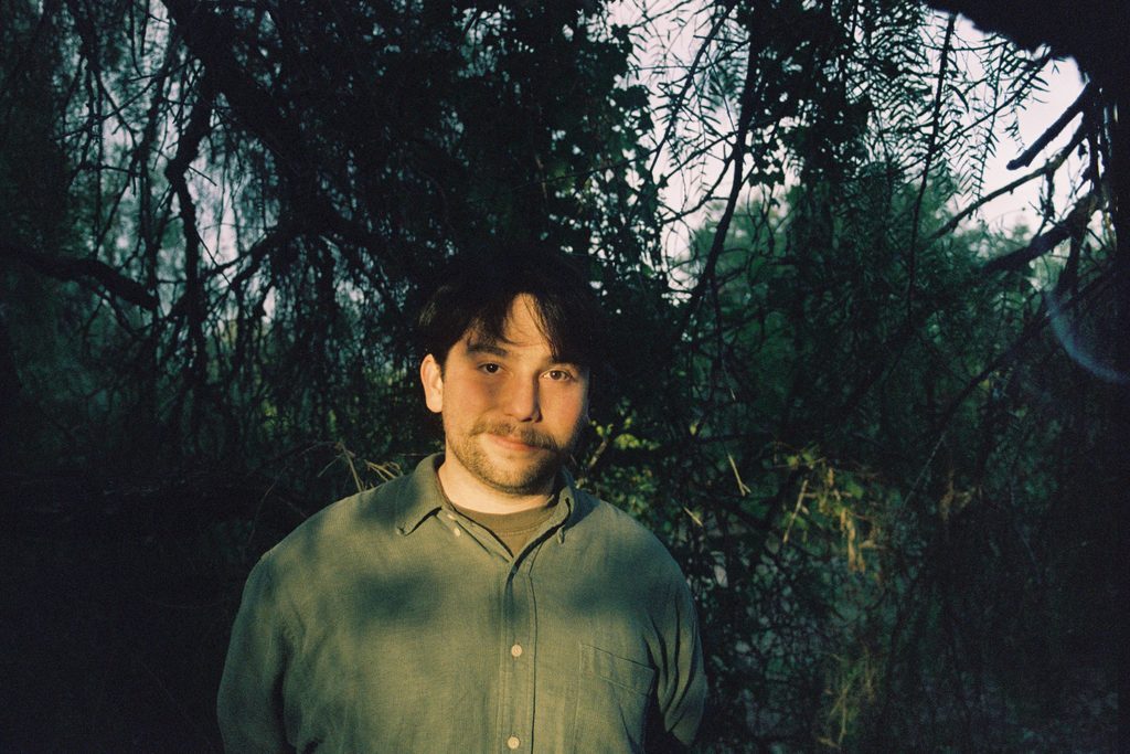 Poet Jacob Herrera Spears stands in a forest. His face is partly in shadow, partly in warm evening light, and he wears a tshirt beneath a green button-down shirt. He has dark brown hair and facial hair.