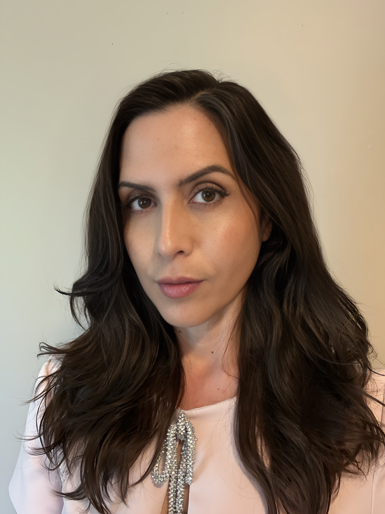 Poet Jenny Sadre-Orafai stands indoors against a light neutral wall. She has long wavy dark brown hair and dark brown eyes. She wears a flowy white blouse with a shimmery silver ribbon tied in a bow.