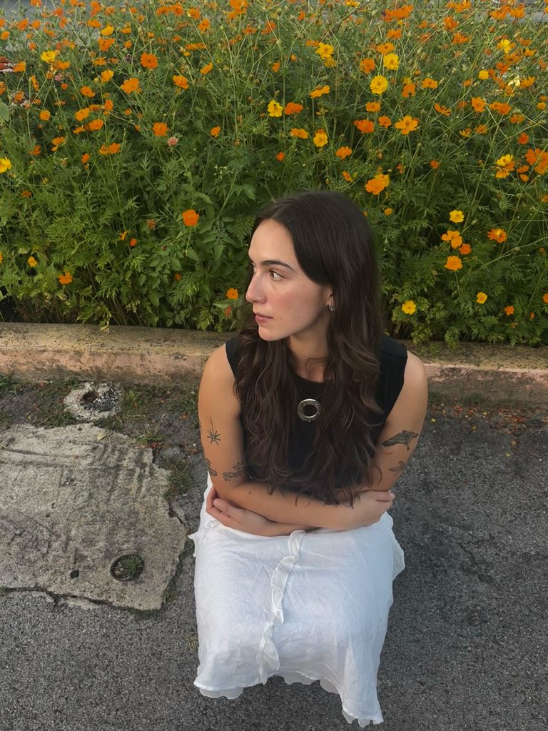 A girl with long, wavy brown hair in a white skirt squats in front of a bush of orange and yellow cosmos flowers. 