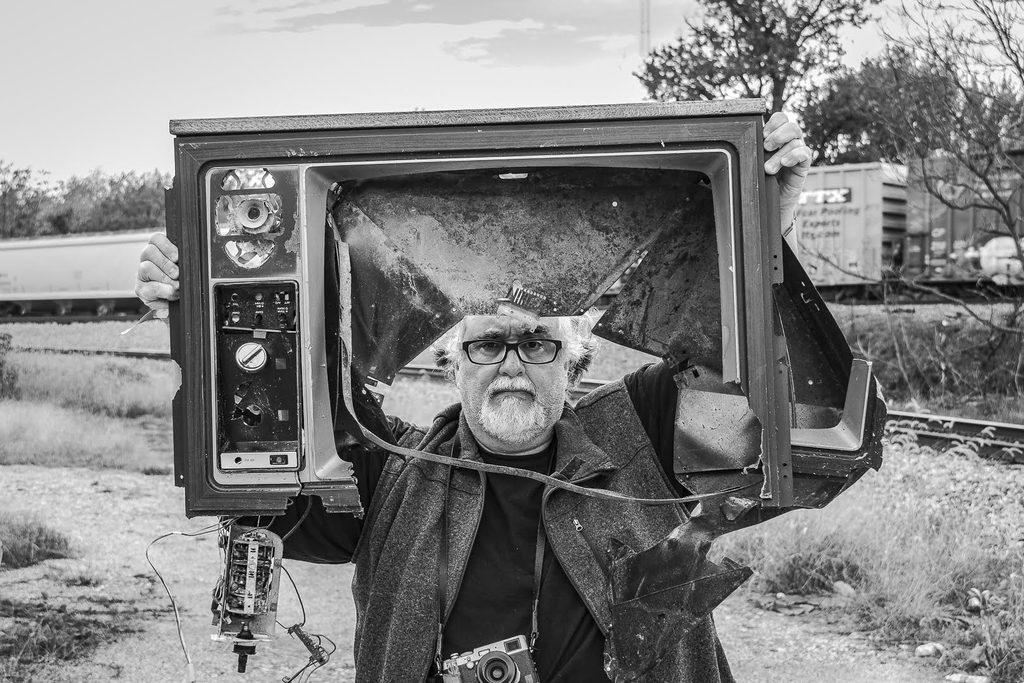 Poet Virgil Suárez stands in a field, holding an broken television. He has white hair and a white beard and wears thick-rimmed glasses, a vest over long sleeves, and an analog film camera around his neck.
